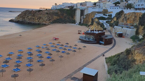 Una playa con sillas azules y sombrillas, una torre de salvavidas y edificios blancos sobre un acantilado.
