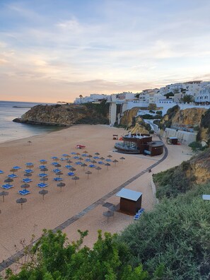 A beach with sun loungers and umbrellas, a lifeguard tower, and a coastal town in the background.
