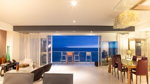 Amplia sala de estar con un gran ventanal que da a un balcón con vista al océano. Hay un comedor con muebles modernos y una araña.