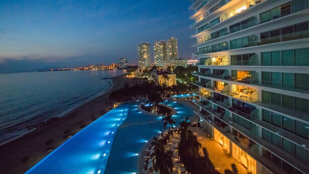 Un hotel frente a la playa con piscina, varios pisos y vista al océano.