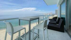 Un balcón con una mesa y sillas de vidrio, con vista a una playa y al océano, y un paisaje urbano distante con edificios.
