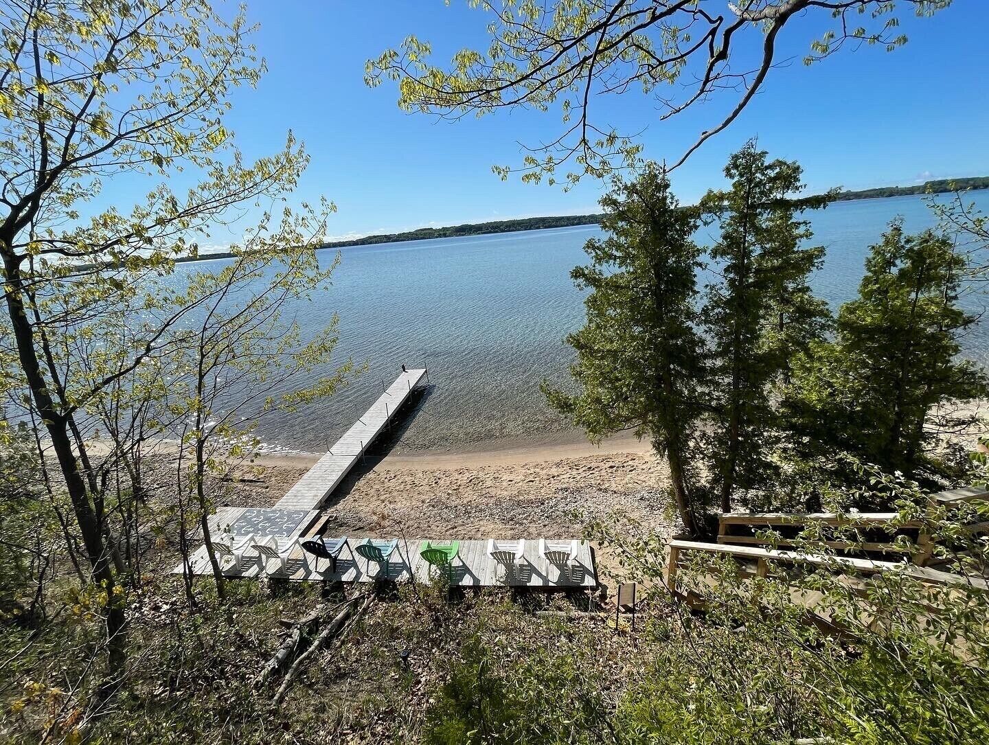 A lakeside view with a wooden dock extending into the water, surrounded by trees and a sandy beach.