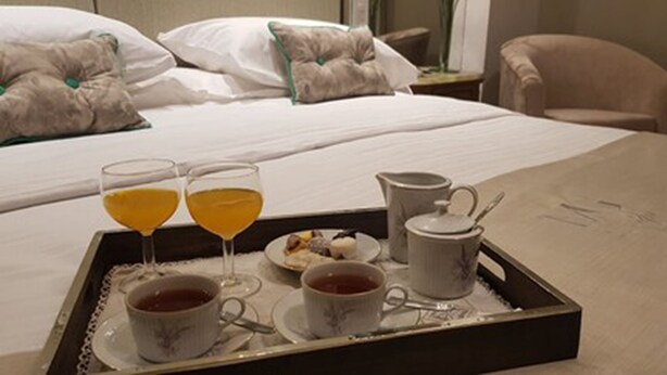 Una bandeja de desayuno bien dispuesta con dos vasos de zumo de naranja, una taza de té y un plato pequeño de pasteles sobre una cama de hotel.