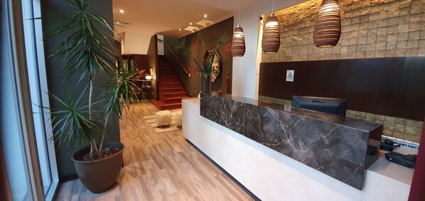 Zona de recepción con mostrador de mármol, plantas en macetas, una escalera y lámparas modernas.