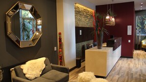 Un área de recepción moderna con mostrador, un sofá gris, un otomano blanco mullido y un espejo hexagonal grande en la pared.