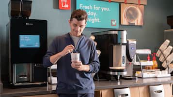 Una persona junto a una máquina de café en un café moderno con un letrero que dice 'Fresh for you. day. night'.