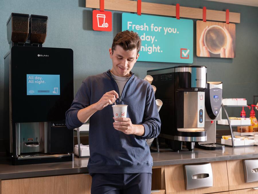 Una persona junto a una máquina de café en un café moderno con un letrero que dice 'Fresh for you. day. night'.
