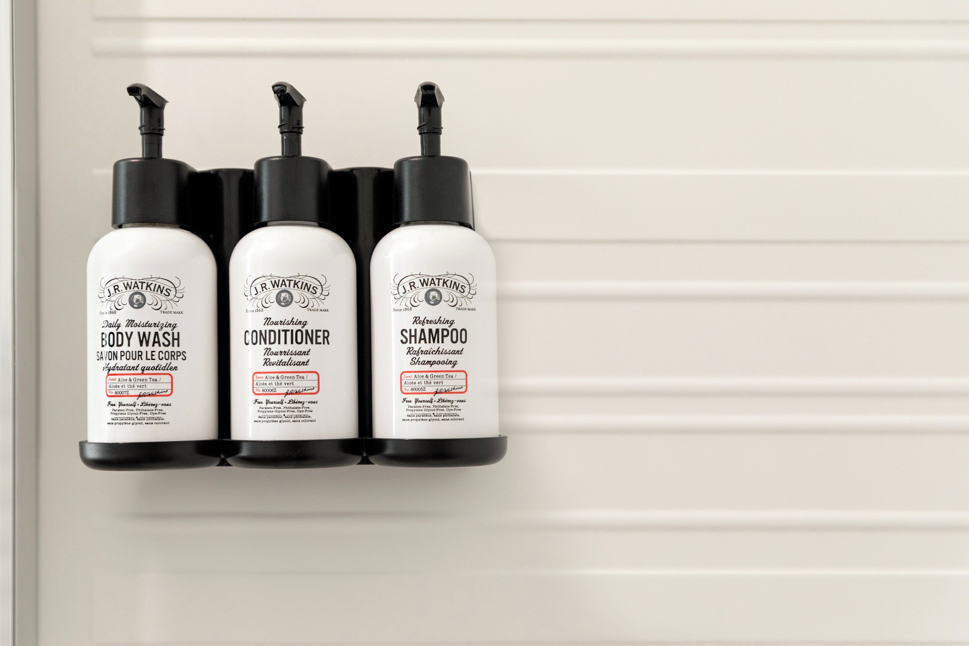 Three J.R. Watkins products: body wash, conditioner, and shampoo, displayed on a white wall.
