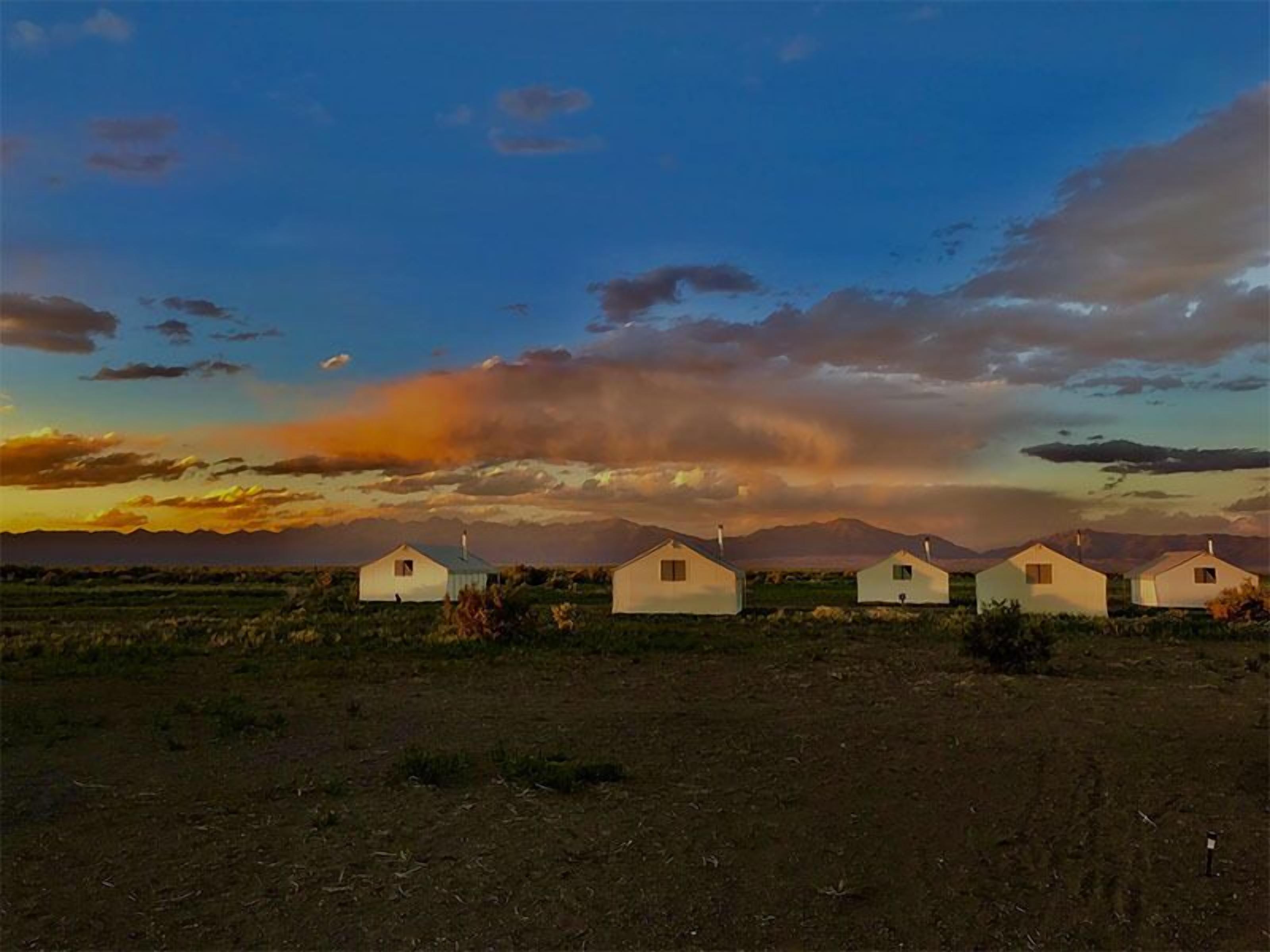 A row of simple, single-story houses with flat roofs, aligned in a straight line across a flat landscape, under a sunset sky with scattered clouds.