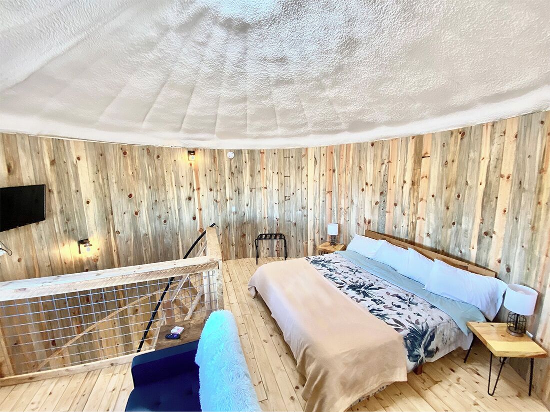 A room with a wooden wall panel design, a bed with a floral bedspread, a small bedside table with a lamp, and a view of the outdoors through a window.