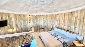 A room with a wooden wall panel design, a bed with a floral bedspread, a small bedside table with a lamp, and a view of the outdoors through a window.