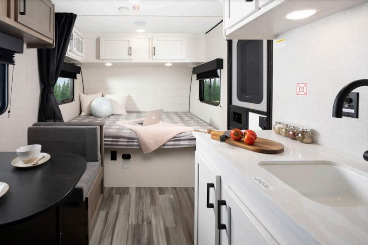 A compact RV interior with a bed, kitchenette, dining area, and a window with a forest view.