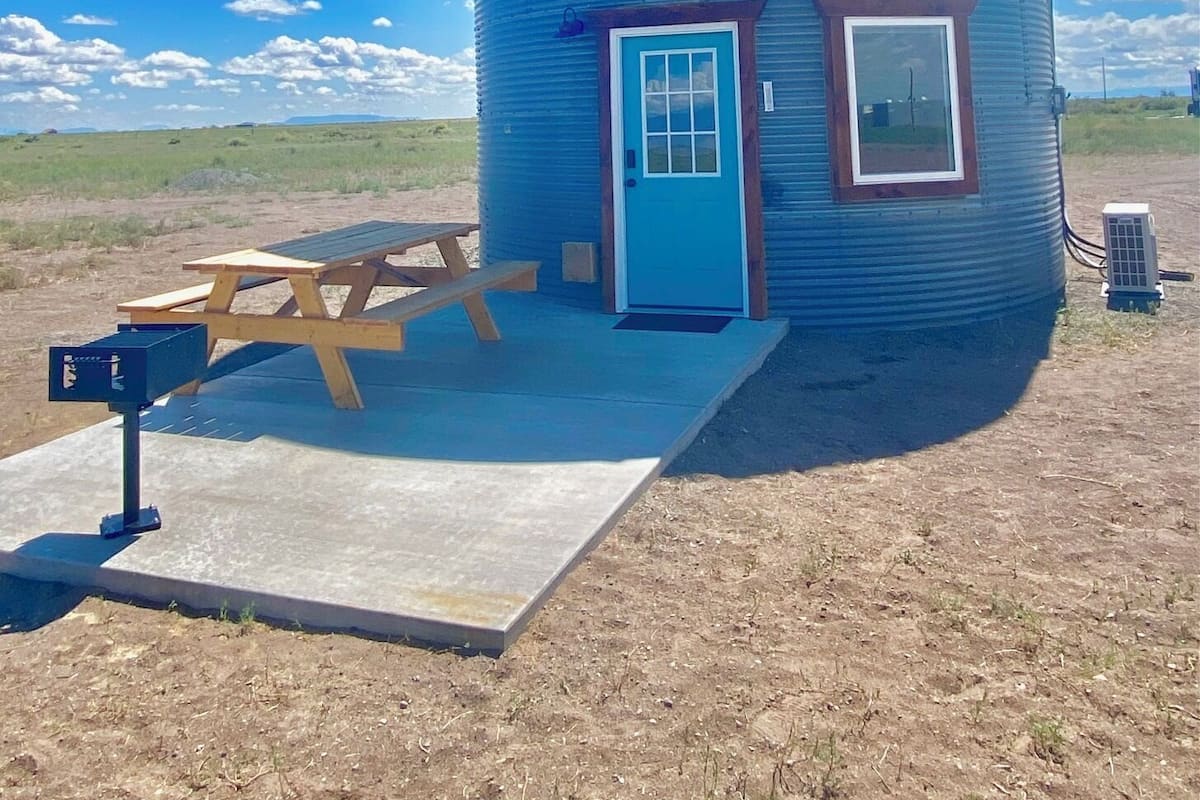 A cylindrical building with a blue door and windows, a picnic table, and a small air conditioning unit.