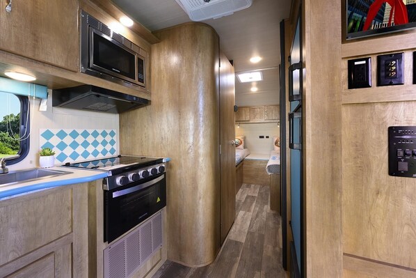 A compact kitchen with a stove, microwave, and sink, and a small sleeping area with a bed and a toilet.