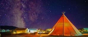 A large, illuminated tent in a rural setting at night, surrounded by small buildings and a starry sky.