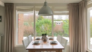 A dining area with a wooden table set for four, surrounded by white chairs, a hanging pendant light, and a view of a garden and buildings outside.