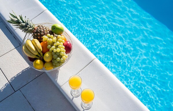 En frugtkurv med bananer, druer, æbler og en ananas ved en swimmingpool.