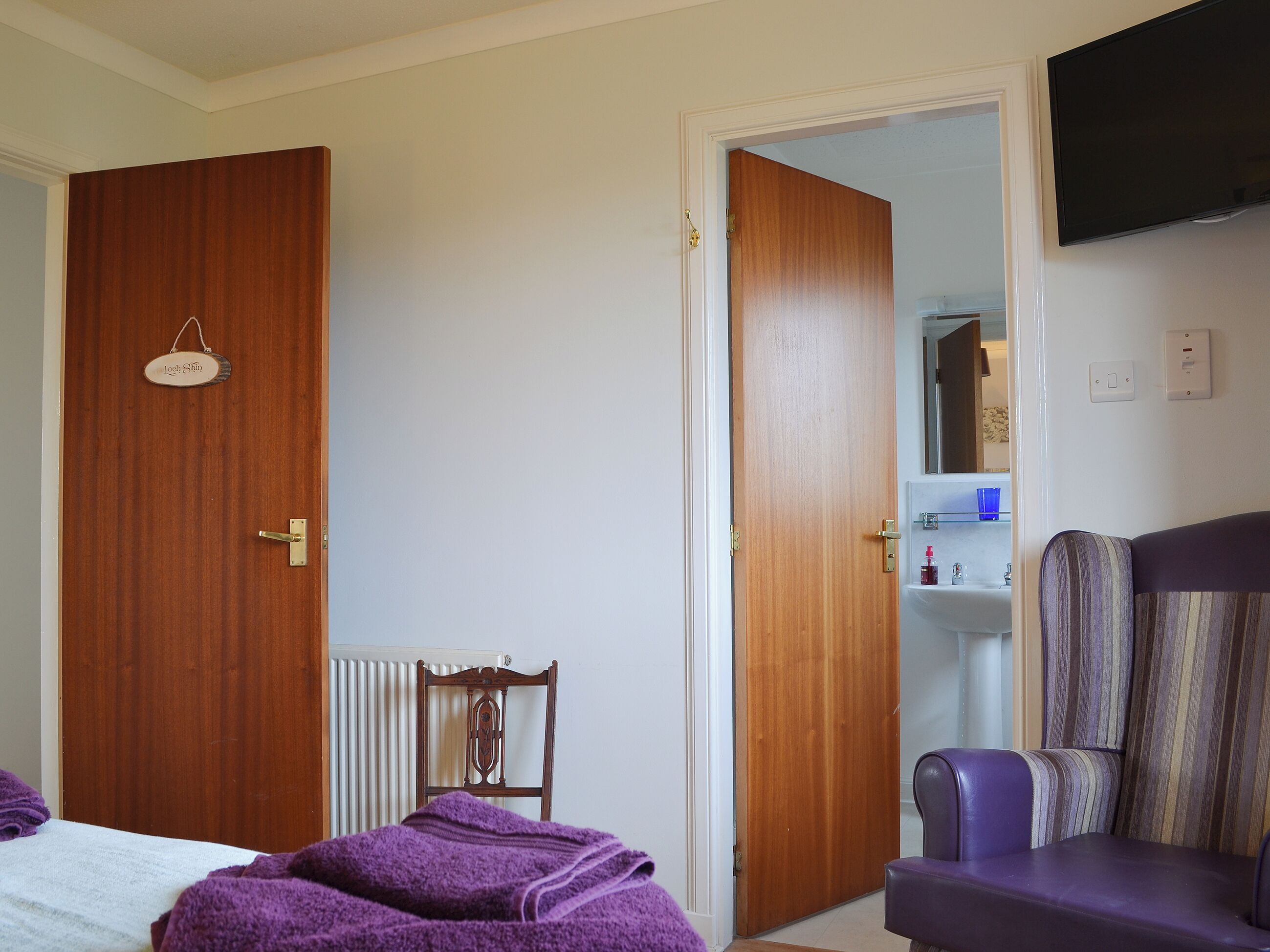 A room with a bed, a chair, a wooden door, and a television mounted on the wall.