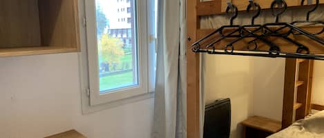 A wooden bunk bed with a metal hanger bar, a small wooden table, and a window with a view of buildings and trees.