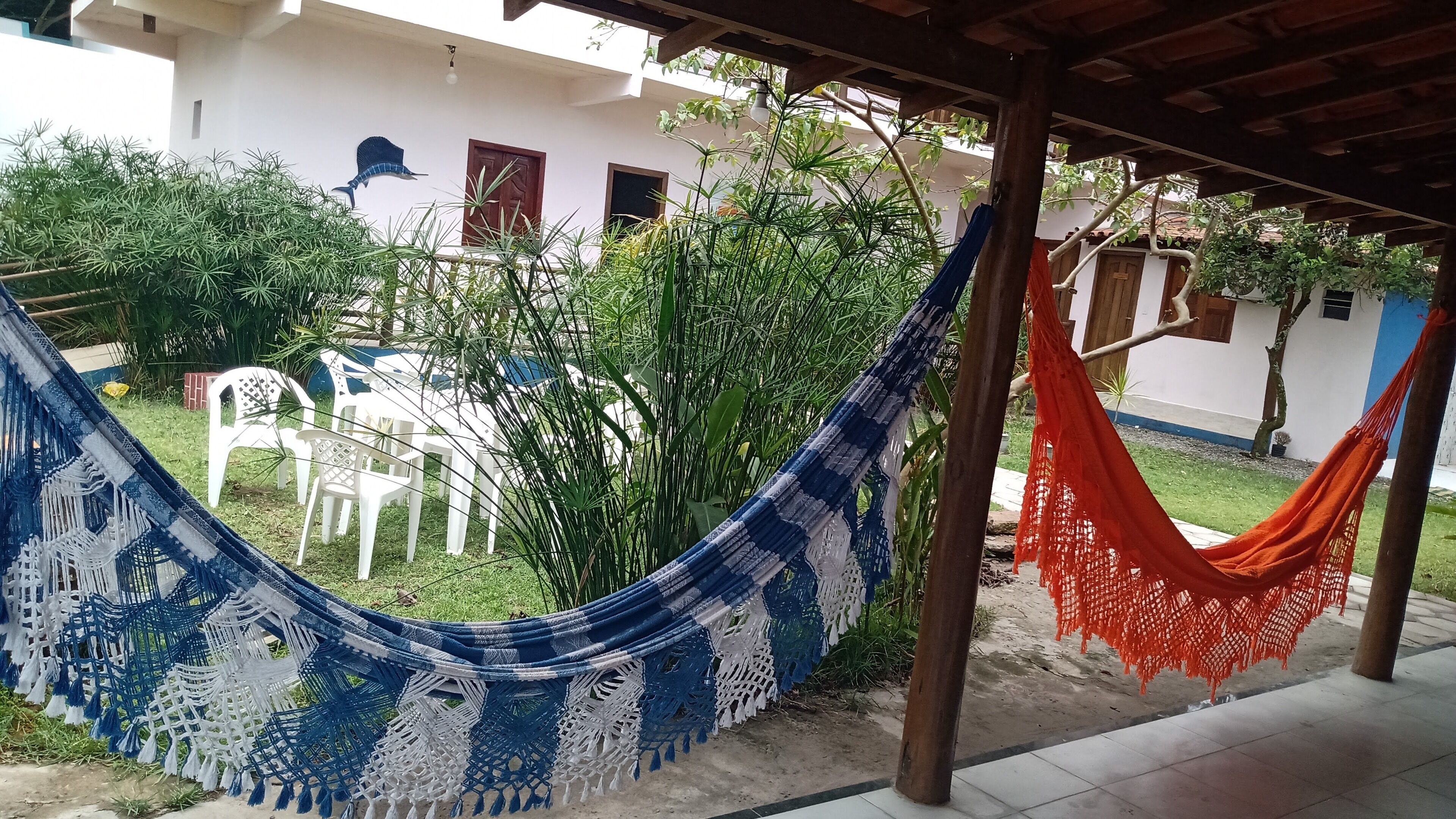 Una hamaca con detalles de macramé, una silla de plástico blanco y una hamaca roja en un espacio exterior cubierto, con un edificio al fondo.