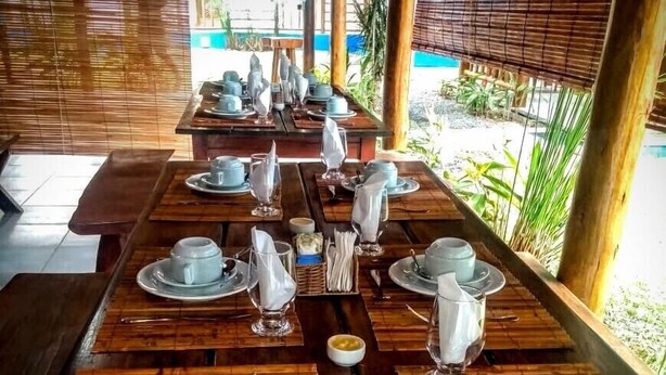 A long wooden table set with white plates, cups, and cutlery, with a view of greenery and a building in the background. - Pousada Pôr do Sol (Maraú)