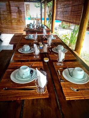 Une longue table en bois, dressée avec de la vaisselle blanche, des tasses et des couverts, avec vue sur un paysage verdoyant et un bâtiment en arrière-plan.