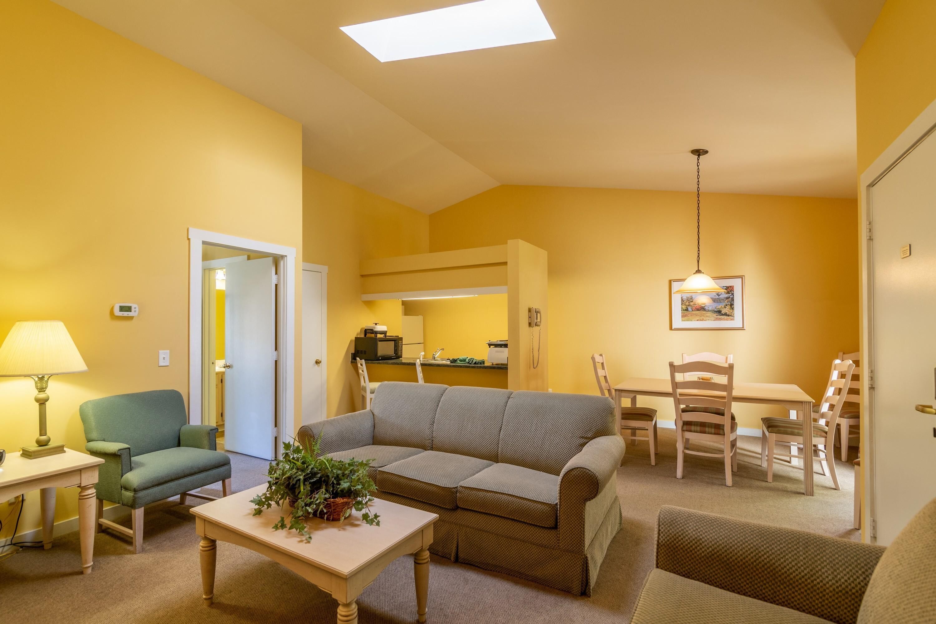 A living room with a sofa, chairs, a table, and a lamp. There is a door leading to a kitchen area.