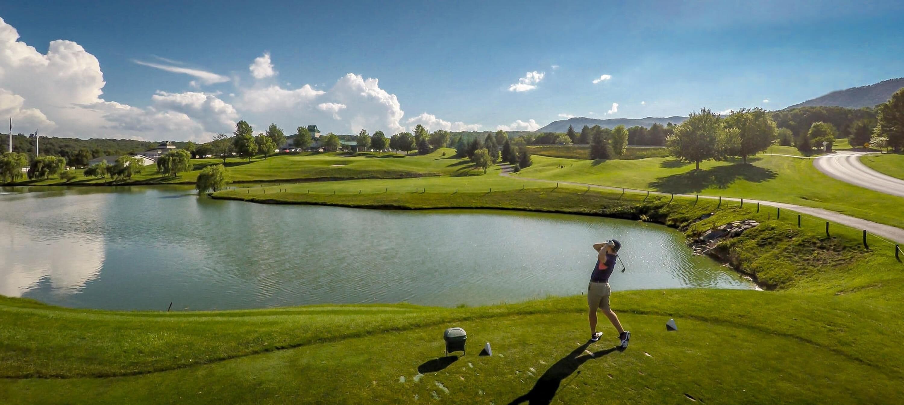 A golf course with a pond, a road, and a building in the background.