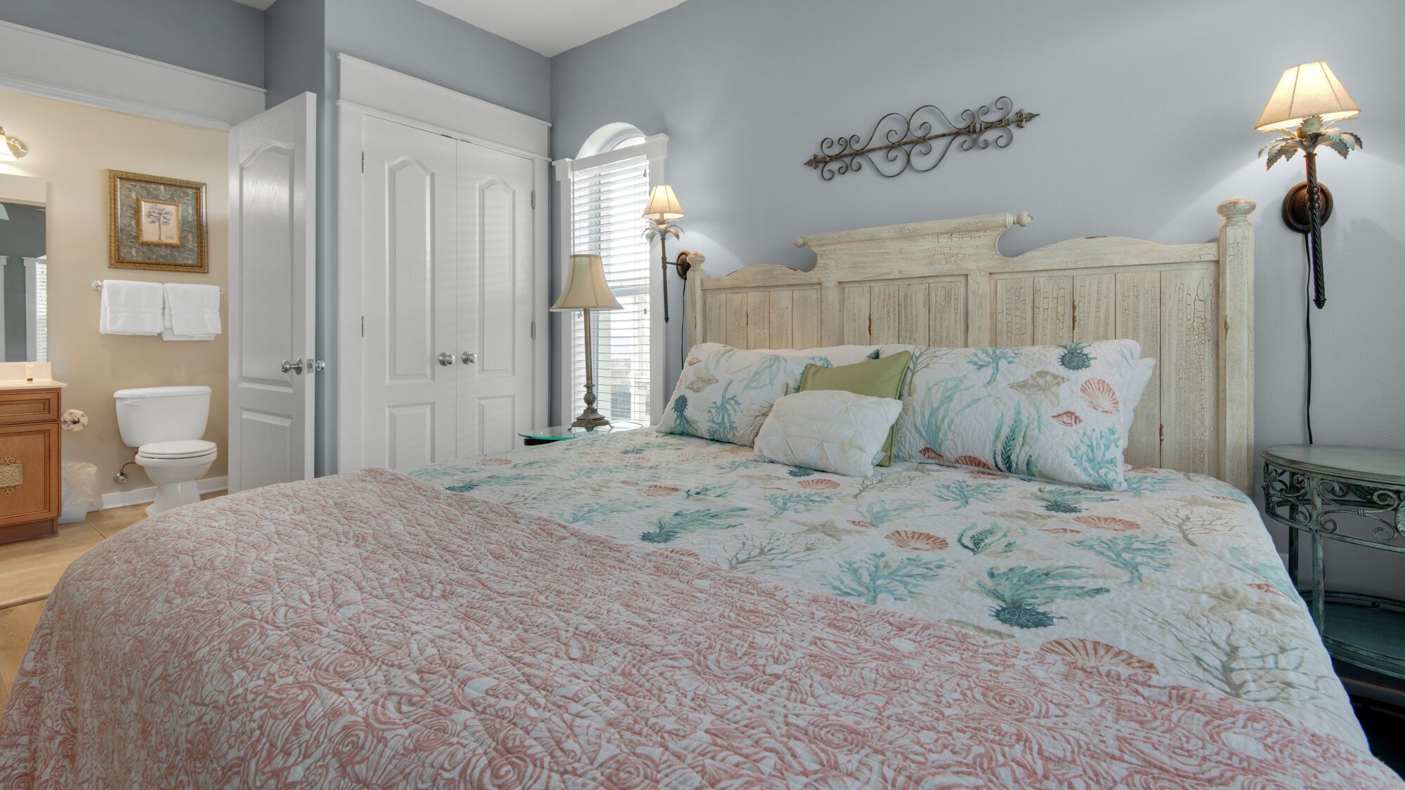 A bedroom with a large bed, a wooden headboard, two bedside lamps, a nightstand, a dresser, and a bathroom with a toilet and towel rack.
