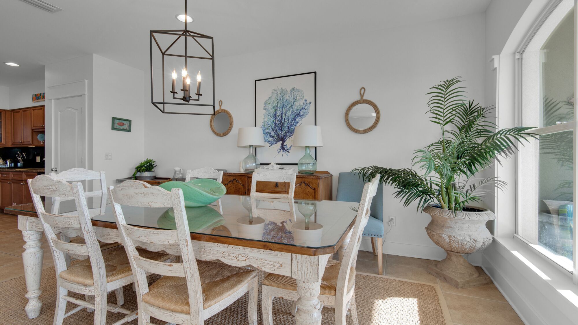 A dining area with a glass table, white distressed chairs, a hanging light fixture, and a large potted plant by the window.