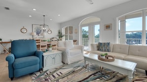 Uma sala de estar ampla com uma poltrona azul, sofá branco e mesa de centro. Janelas grandes oferecem vista para o exterior.