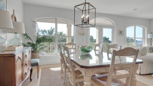 Uma ampla área de jantar com mesa de vidro, cadeiras brancas e vista para o exterior através de grandes janelas.