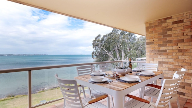 A dining area with a table set for four, overlooking a beach and the sea. - Pelican Sands, 3/83 Soldiers Point Rd - stunning waterfront unit, magical views & air conditioning (Soldiers Point)