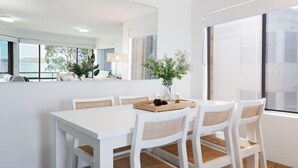 A modern dining area with a white table and chairs, a vase with flowers, and a window with a view of the outdoors. - Pelican Sands, 3/83 Soldiers Point Rd - stunning waterfront unit, magical views & air conditioning (Soldiers Point)
