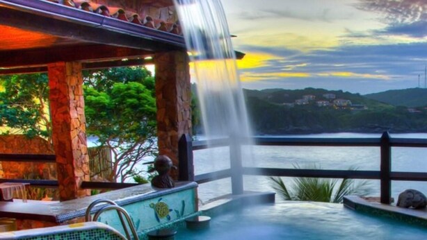 Zona de piscina con cascada, sillas de descanso y vista a un cuerpo de agua y colinas al fondo.