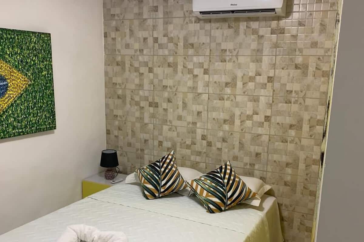 A hotel room with a bed, a wall-mounted air conditioner, a decorative wall art, and a tiled wall.