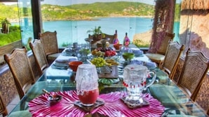 Mesa de comedor preparada con vistas a un lago y colinas, rodeada de sillas de mimbre.