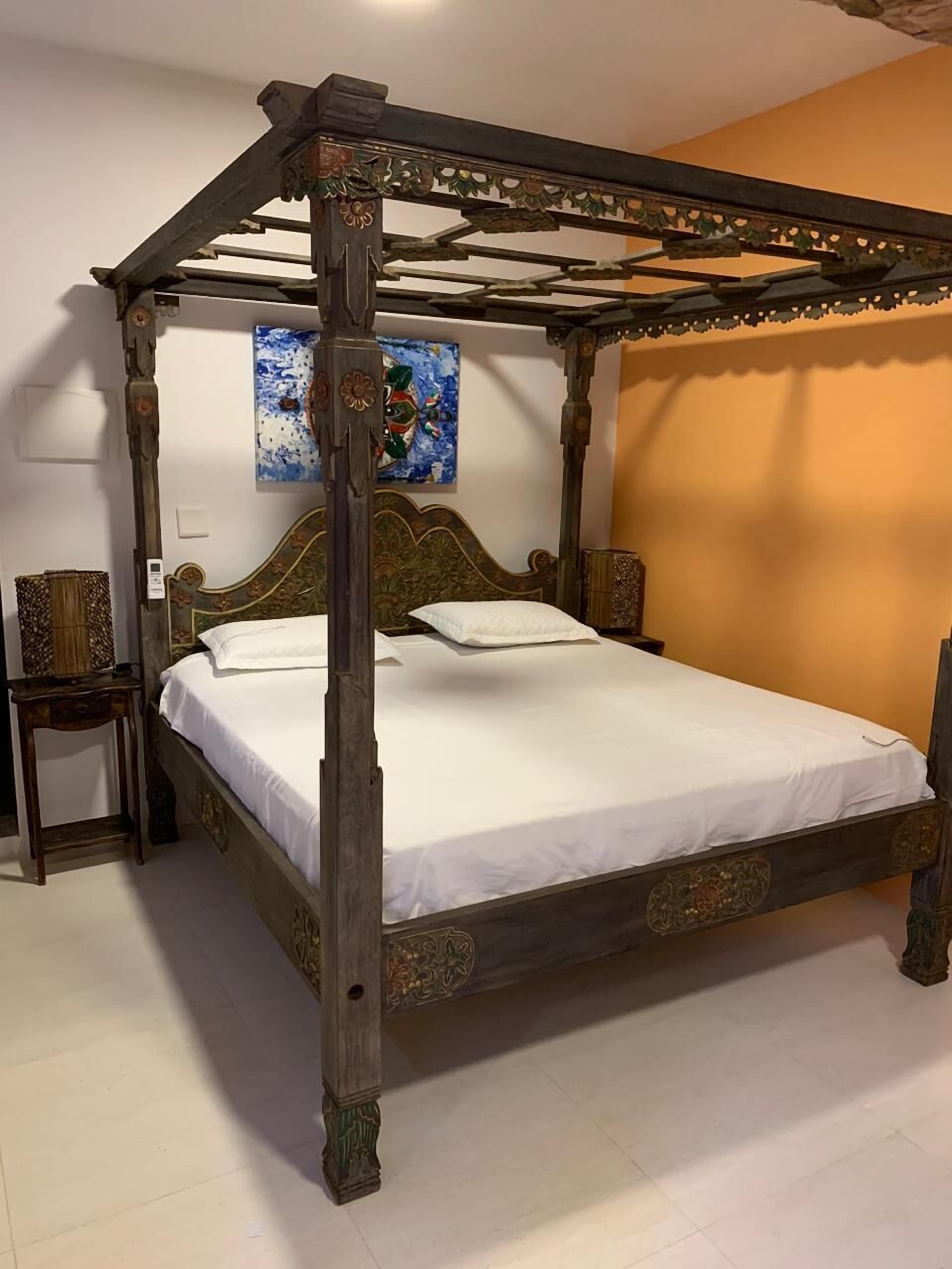 A four-poster bed with a wooden frame and intricate carvings, a bedside table, and a wall-mounted lamp.