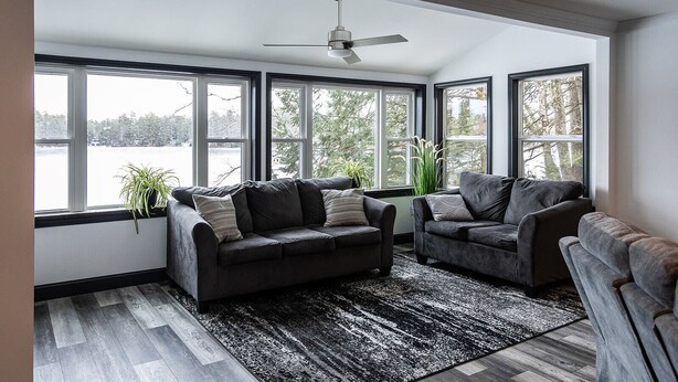A modern living room with a dark leather sofa, a smaller armchair, and a large window with a view of a lake. - Luxurious Lakeside Retreat on Pequawket Lake (Limington)