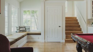 Een kamer met houten vloer, een pooltafel, een bank met kussens, een deur en een trap.