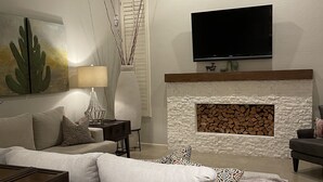 Una sala de estar con chimenea, un televisor de pantalla plana montado en la pared, un ventilador de techo y una gran obra de arte con un cactus.