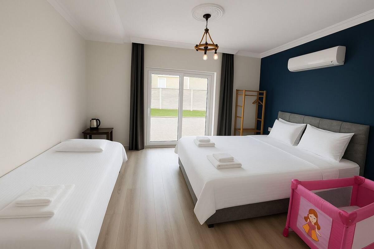A hotel room with two beds, a small table, a chair, a ceiling light, and a view of the outside.