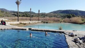 A clear pool with two people swimming, surrounded by a natural landscape with hills and trees. - Dalaman Airport Guesthouse (Dalaman)