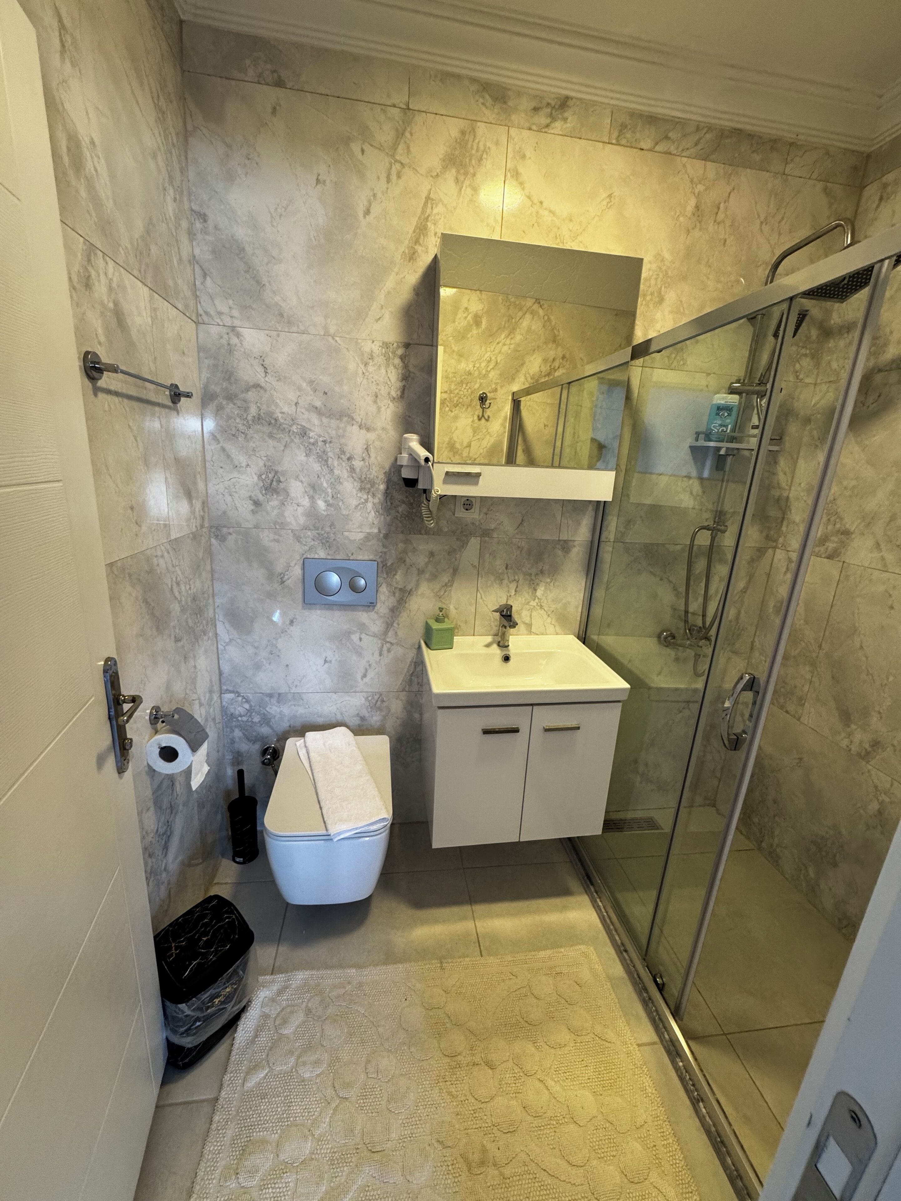 Un baño con cabina de ducha de vidrio, un lavabo blanco, un espejo sobre el lavabo y un inodoro con una toalla encima.