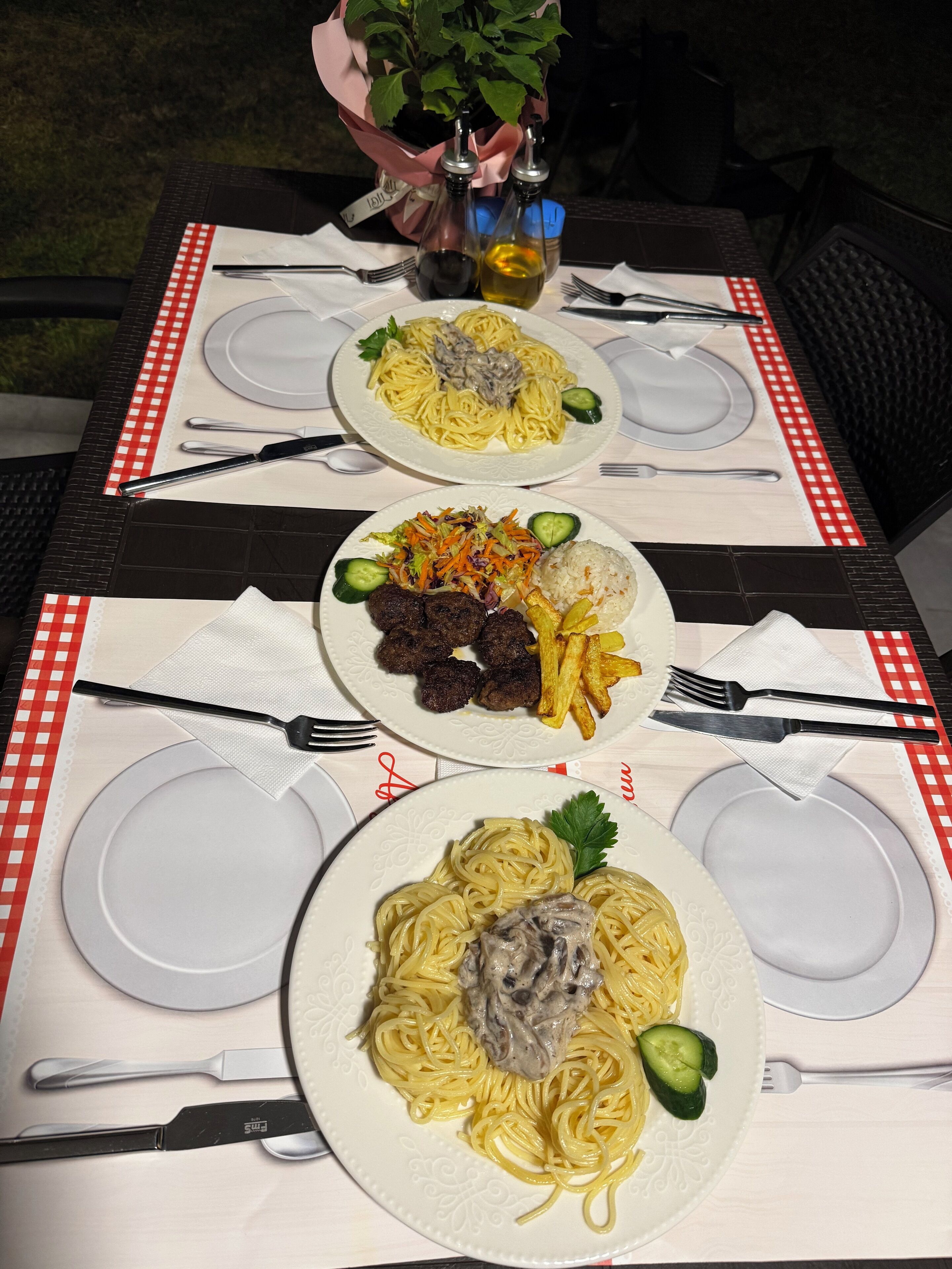 Ein Tisch für drei Personen, gedeckt mit Spaghetti, Fleischbällchen und Pommes, sowie Gewürzen und Besteck.
