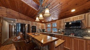 A spacious kitchen with wooden cabinets, a large island with bar stools, stainless steel appliances, and a stone backsplash. - Log home mins from Lake George, w/ heated salt water pool and hot tub! (Warrensburg)