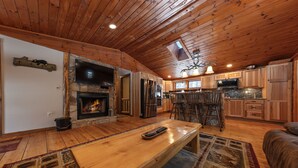 A spacious living area with a wooden ceiling, a stone fireplace, and a large wooden dining table. - Log home mins from Lake George, w/ heated salt water pool and hot tub! (Warrensburg)