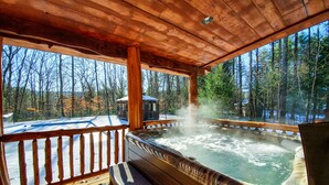 A wooden deck with a hot tub, wooden benches, and a view of a forested area. - Log home mins from Lake George, w/ heated salt water pool and hot tub! (Warrensburg)
