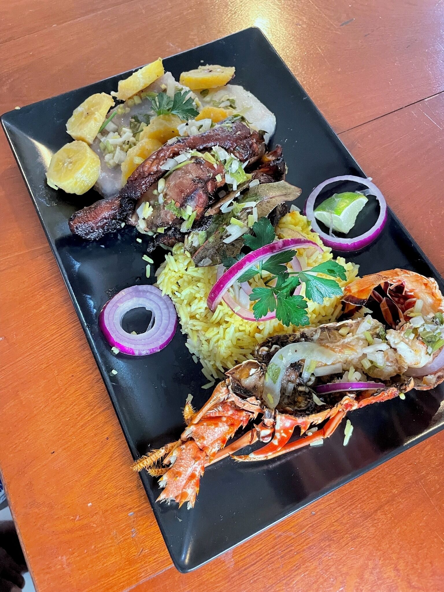 A black rectangular plate with a variety of seafood including a whole crab, shrimp, and pieces of fish, accompanied by yellow rice, potato slices, and garnishes like parsley and red onion rings.