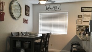 Un comedor con una mesa, sillas y una ventana con persianas. Hay adornos en la pared y un ventilador de techo.